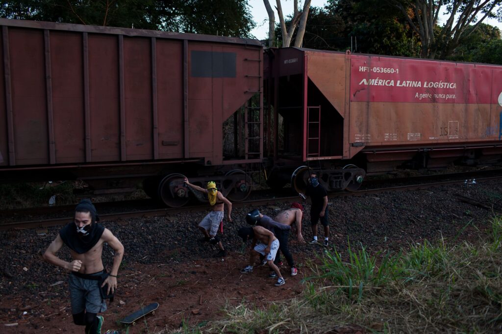 Grupo de homens aparece sem camisa ao lado de um trem de carga. Eles estão em movimento. 