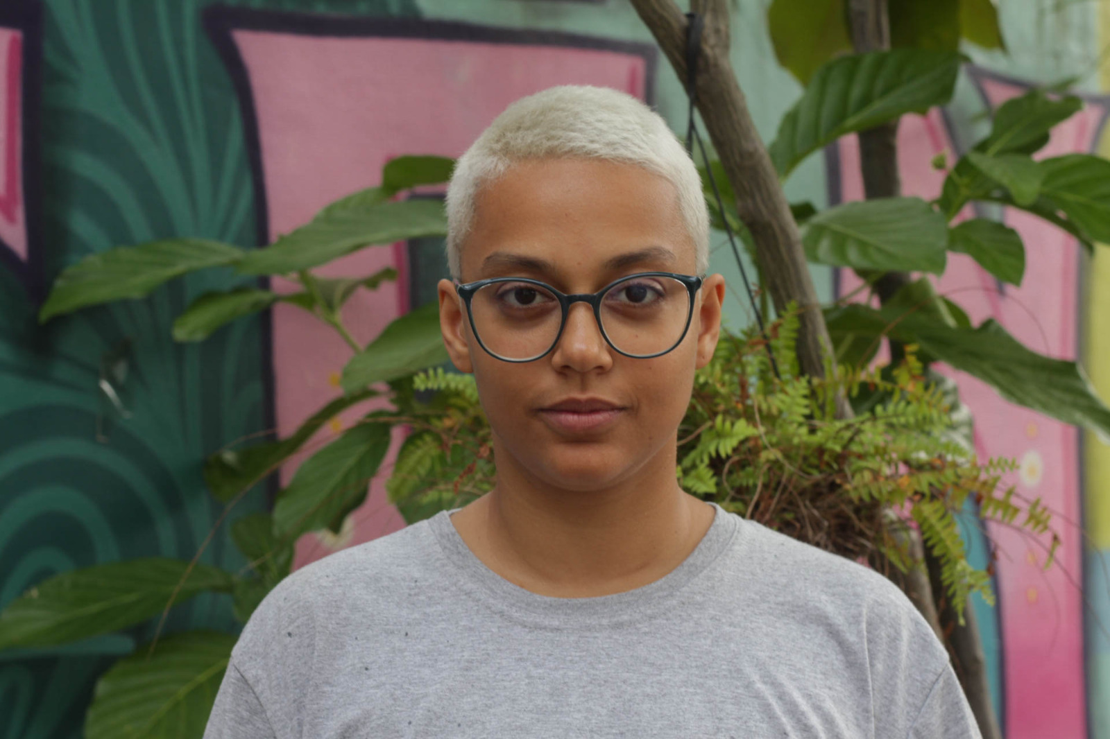 foto da artista. Cabelos curtos com fios claros. Óculos de grau com armação preta, veste camisa cinza e o fundo da foto tem paredes rosas e verdes, além de plantas na decoração.